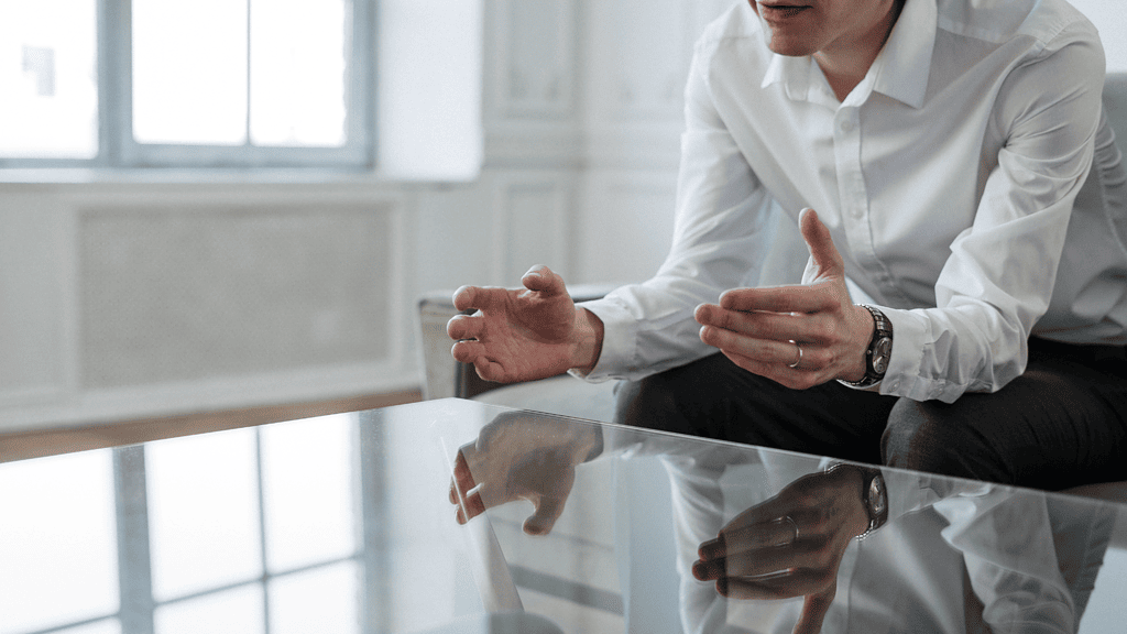 man in a white shirt seated at a glass table leaning forward and gesturing with both hands