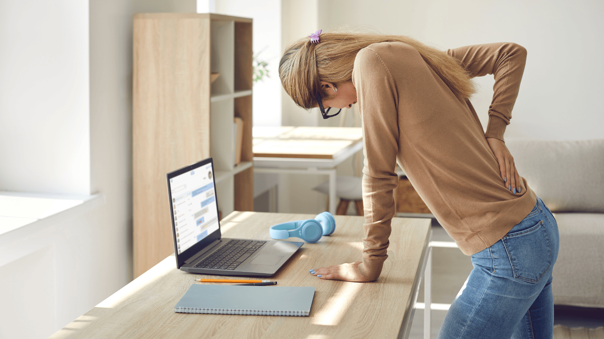 a pained woman leans on her desk with a hand on her lower back