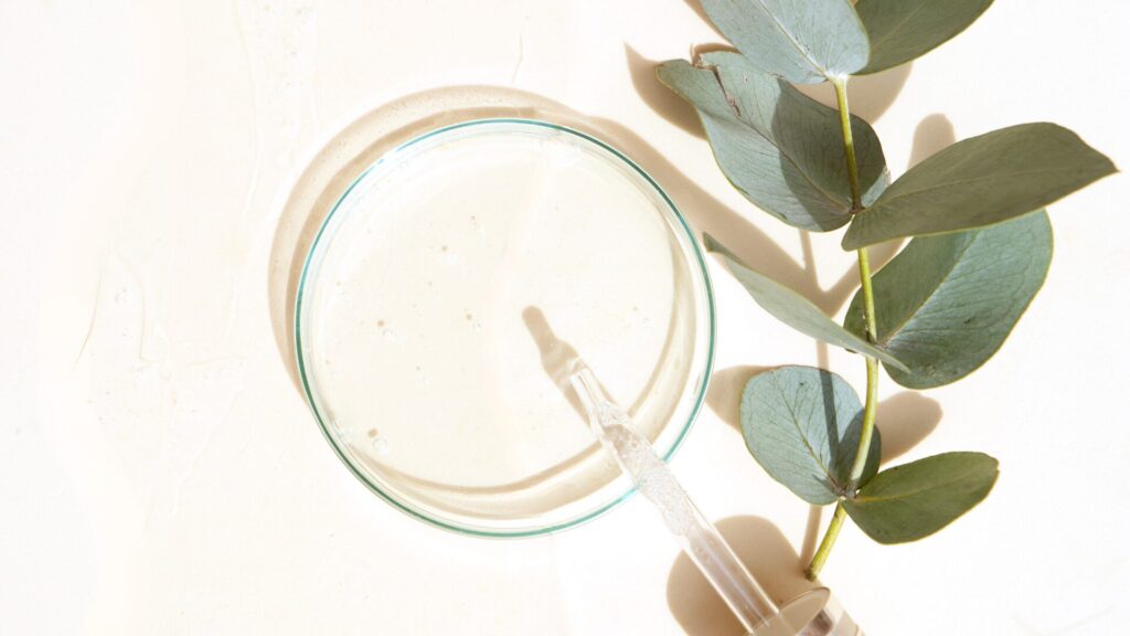 An overhead photo of an eye dropper resting on a petri dish with a eucalyptus brand beside it
