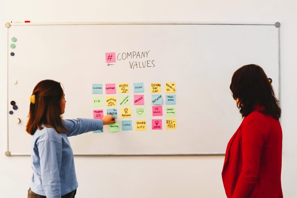 two women in front of whiteboard that has sticky notes with company values on them